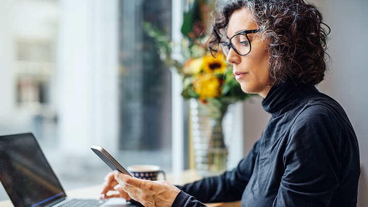 Frau schaut aufs Handy, während sie am Laptop arbeitet