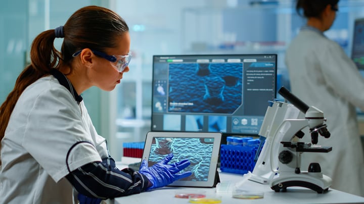 Scientist wearing goggles in lab at desk with iPad, computer and microscope