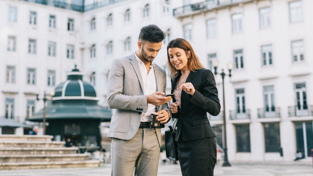 Two business professionals smiling and looking at a smartphone outdoors in a city square.