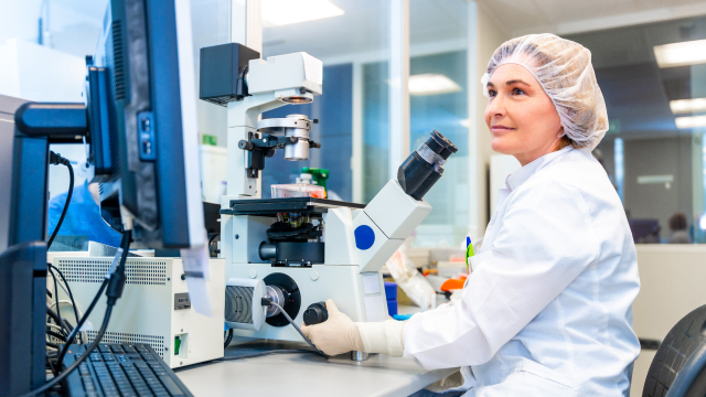 Lab technician sitting at a microscope looking up at monitor