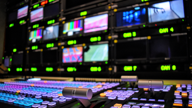 A professional broadcast control room with multiple monitors and a colorful video switching console
