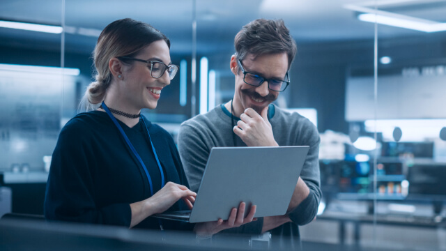 Two coworkers wearing ID badges and glasses review information on a tablet together inside a modern office workspace, smiling as they discuss.