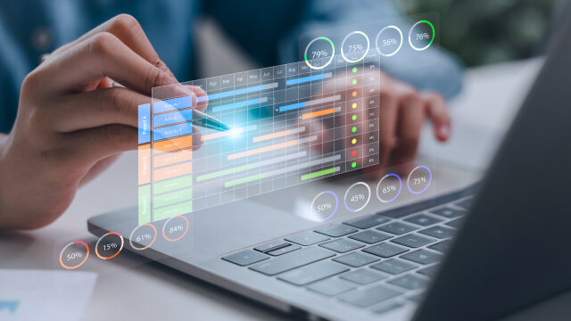 Close-up of a person using a stylus on a transparent digital dashboard overlay above a laptop keyboard, showing charts, progress bars, and data metrics.