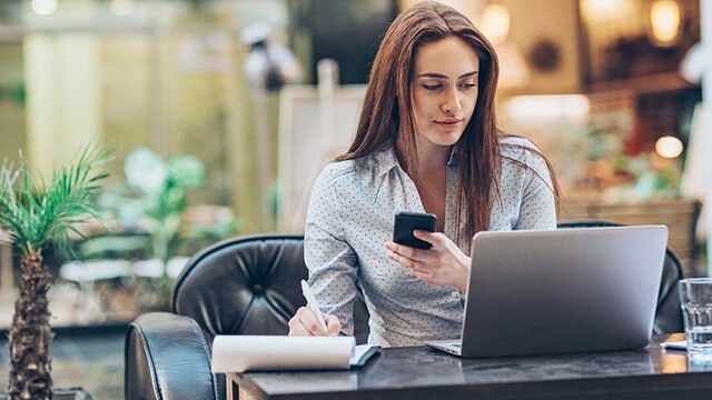 Woman at desk with laptop and phone in hand