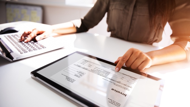 Person reviewing a digital invoice on a tablet while working at a desk with a laptop