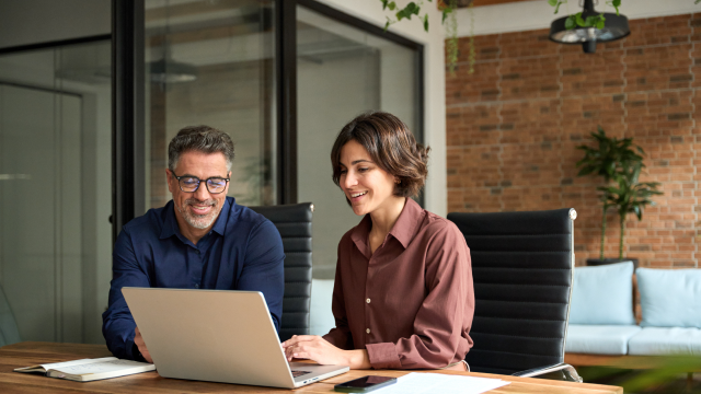 Two people smiling while looking at a laptop together in a modern office