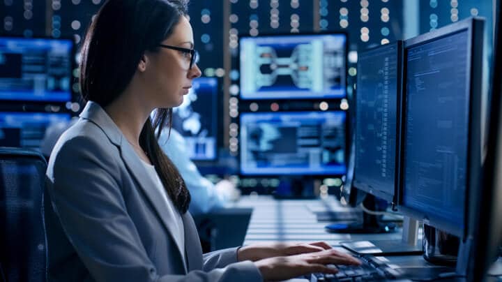 Mujer con gafas en el escritorio escribiendo en el teclado mirando a dos monitores